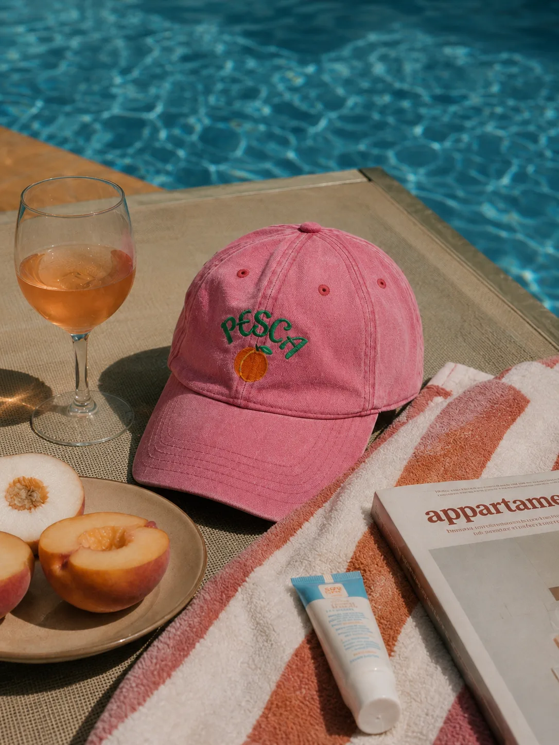 Pink Pesca cap poolside still life with peaches and a glass of rosé