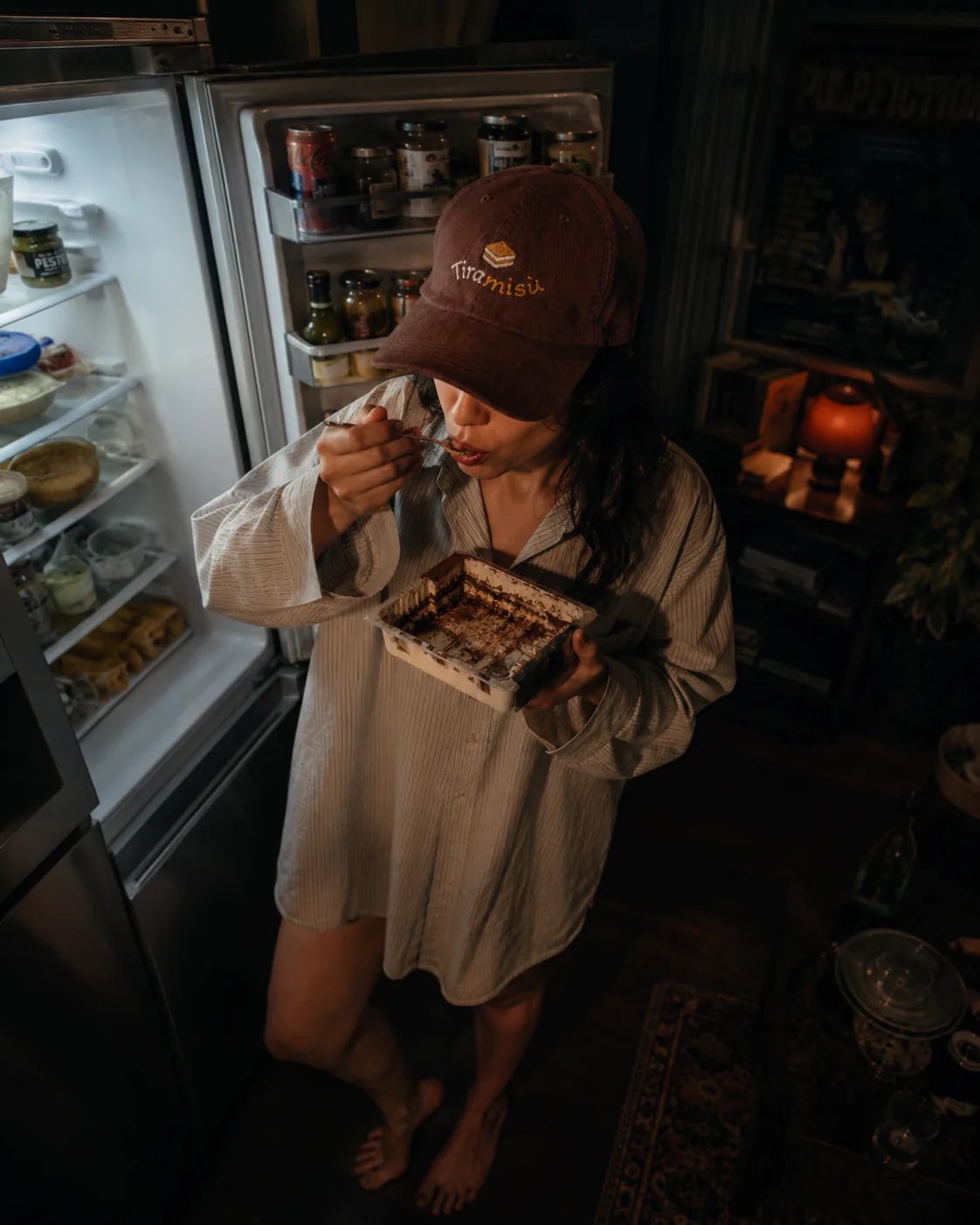 Woman wearing the brown Tiramisù cap, eating tiramisù by an open fridge